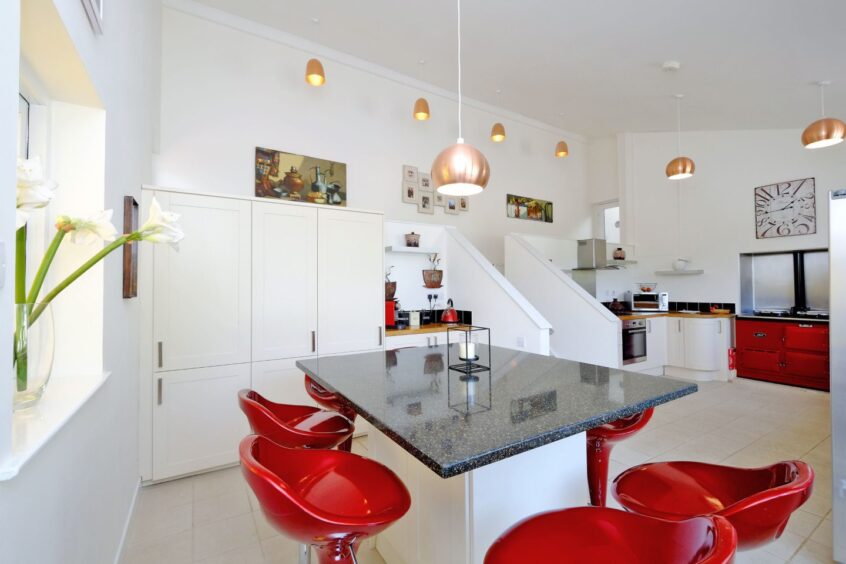 The kitchen with white walls, floors and cupboards, black speckled counter tops and red accents. There is a breakfast island in the centre of the room with sleek red bar stools