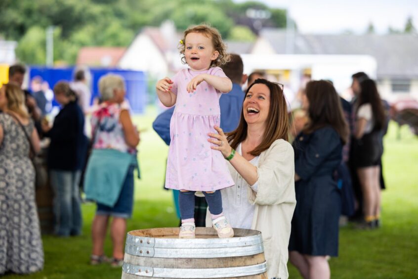 A young girl in a pink dress stands on a barrel with a relative holding her from behind.