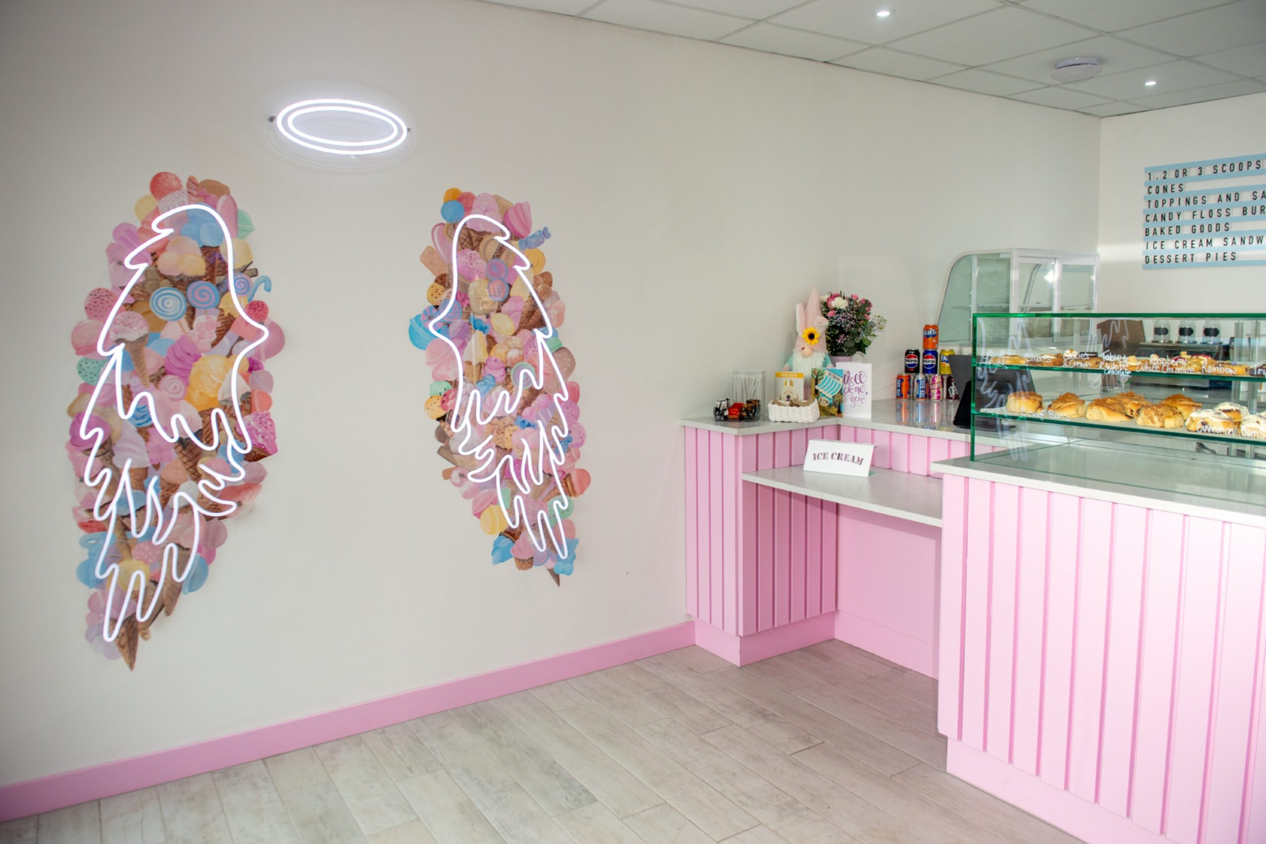 The interior of Lolly's Ice Cream in Portlethen, with pastel colours and neon wings on the wall next to the counter for customers to take photos with