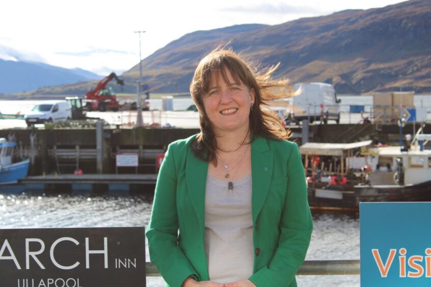 Maree Todd, dressed in a green blazer jacket, standing with Ullapool harbour in the background.