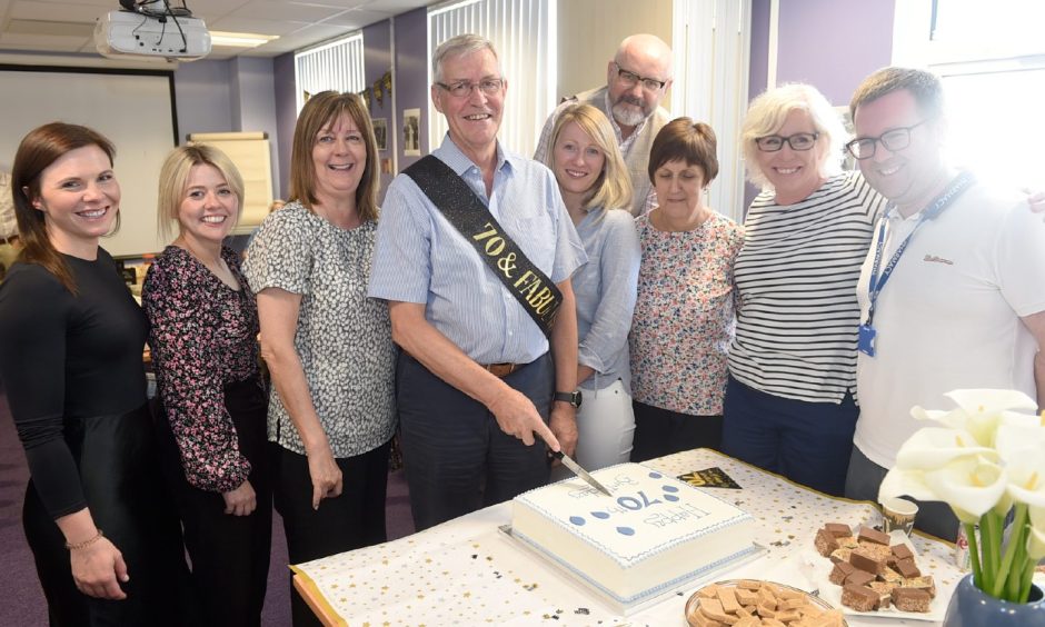 Dr Ron Stewart surrounded by colleagues while cutting 70th birthday cake. 