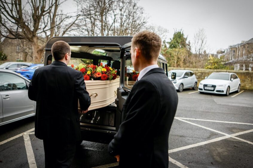 Funeral directors taking a light brown coffin out of the rear of a Land Rover Hearse. 