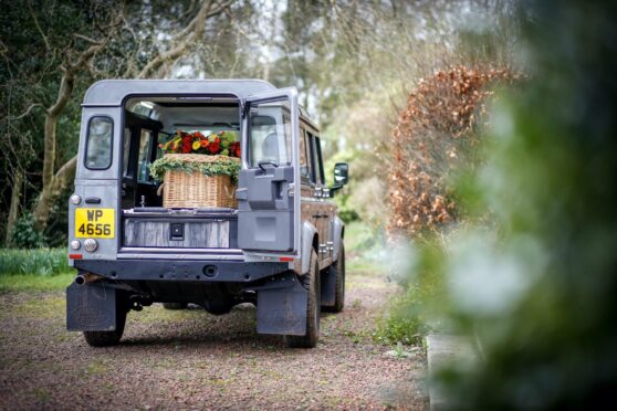 Silver Land Rover Defender hearse with a coffin the back with the door open in woodland.