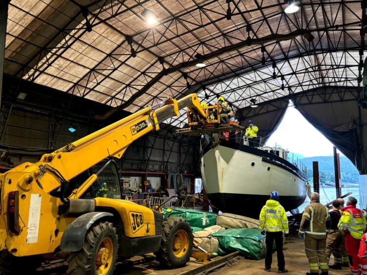 A yellow crane at Mallaig Boatyard with a wooden pallet carrying firefighters from the boat inside the shed.