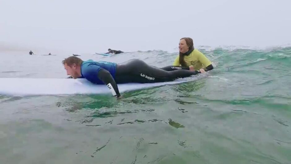 Iona, wearing a yellow top, pictured in the water with a man on a surf board in front of her.