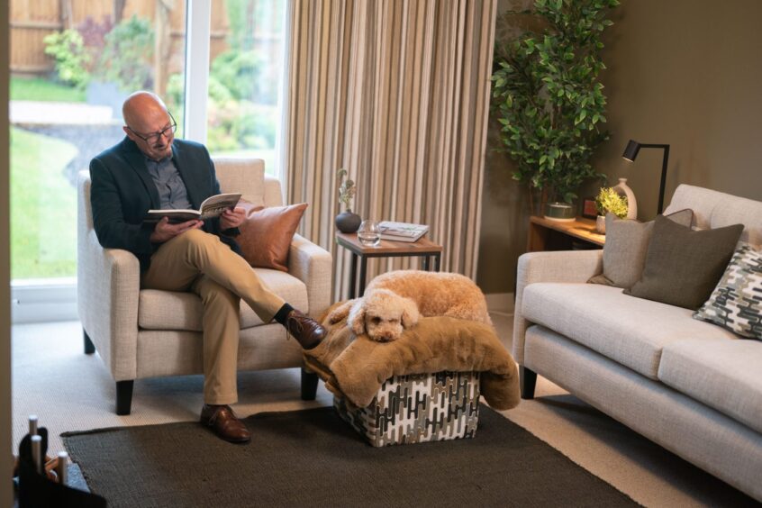 man sitting in a cosy living room reading a book with a dog lying down on an ottoman, an example of the lifestyle inside Scotia Homes' Dalfaber development