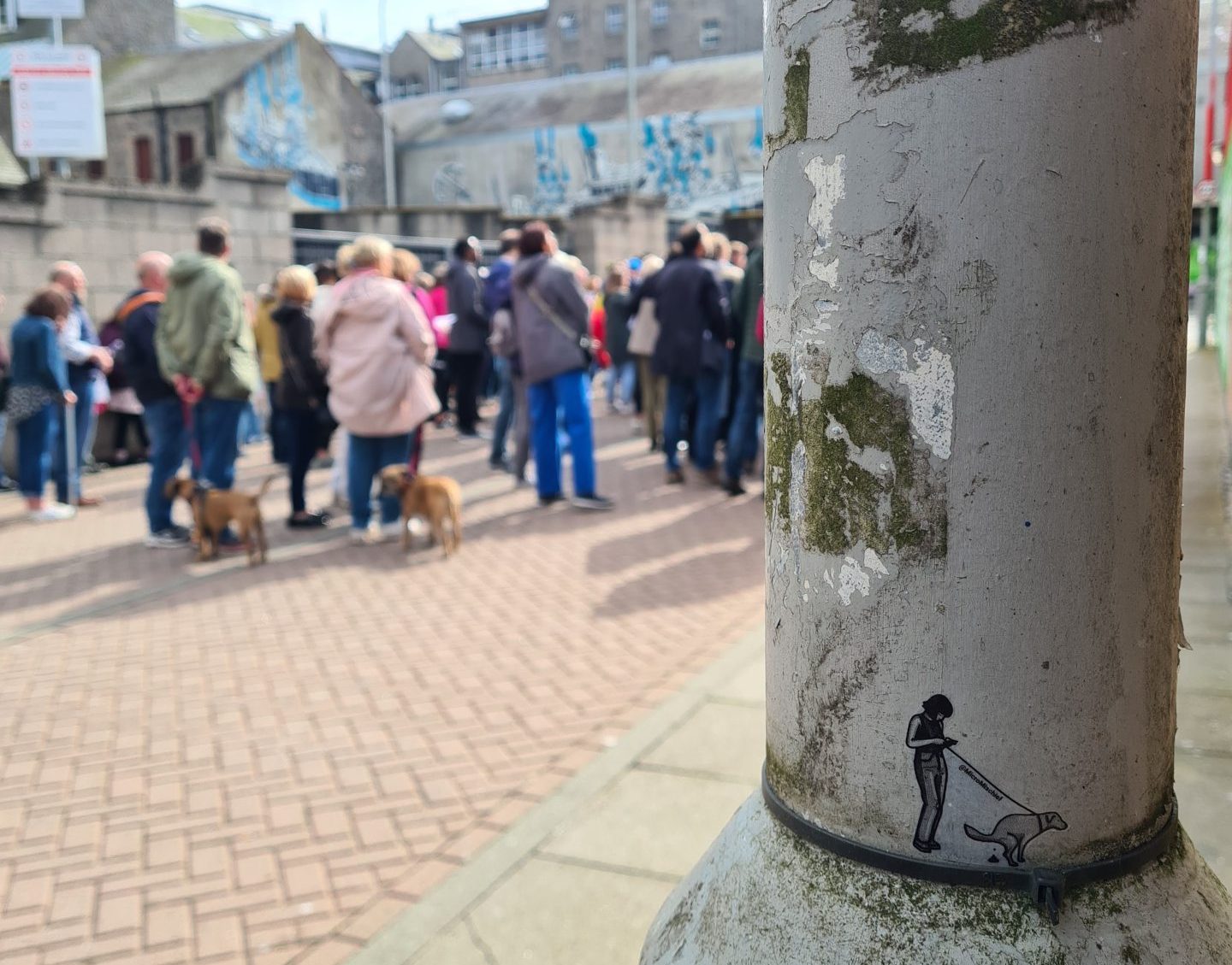 A sticker of a person with a dog on a lead as people with dogs on leads look at Nuart murals in Aberdeen. Image: MicroMischief.