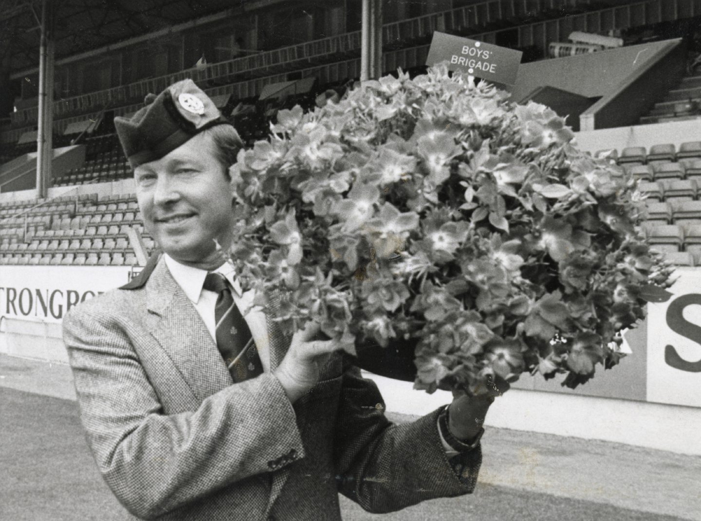 Image shows football manager Alex Ferguson standing on the pitch at Pittodrie, holding a large bunch of roses for the Boys' Brigade centenary in 1983.