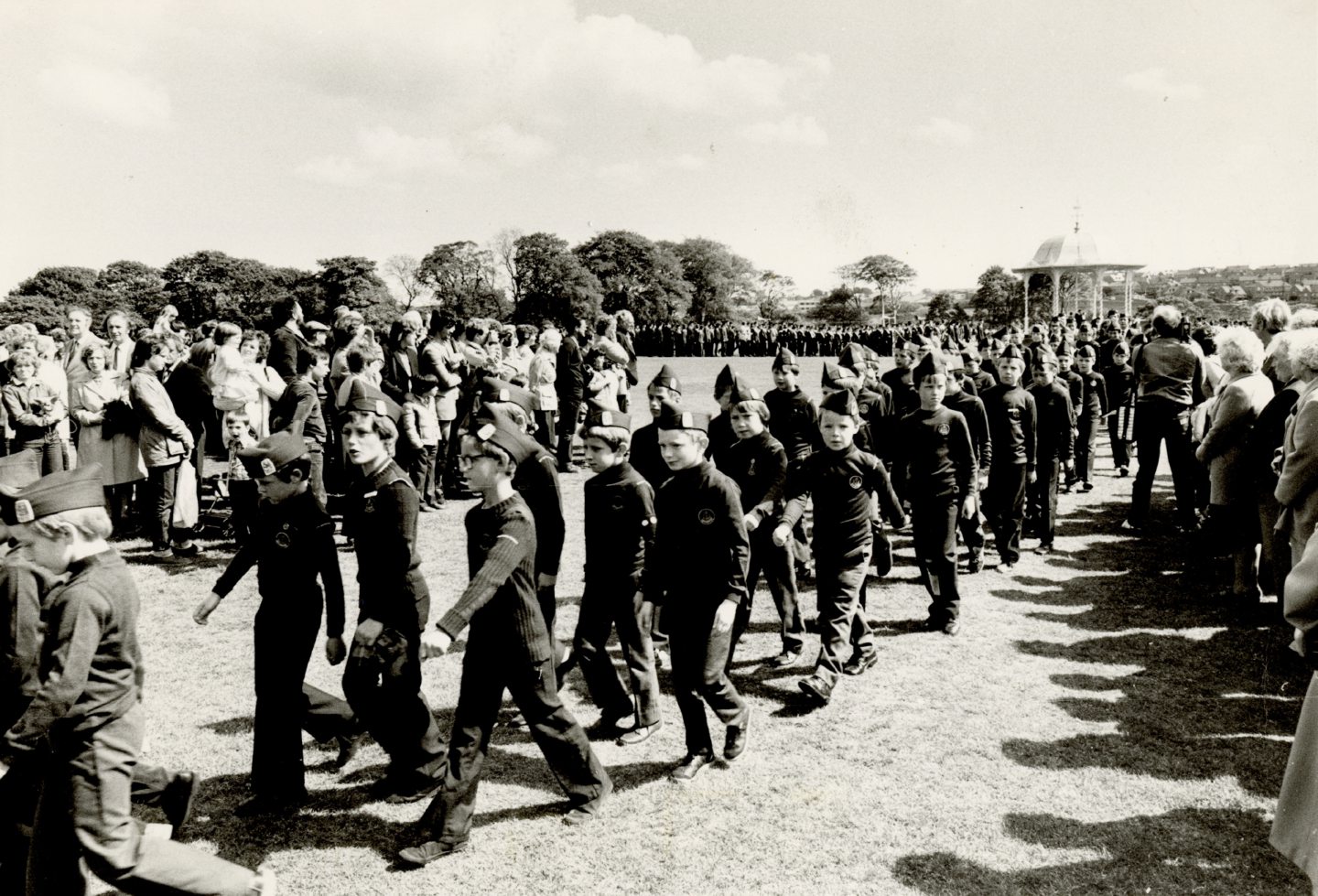 Image shows a large group of boys in their uniforms, marching in the park. There is a crowd of onlookers watching as the boys pass