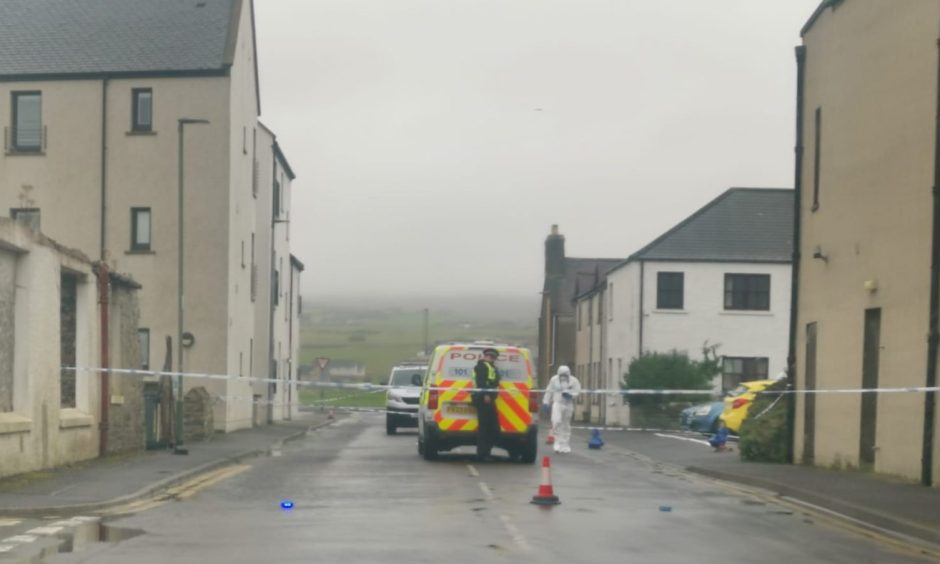 Forensic officer next to police car and officer behind police tape on a street in Orkney.