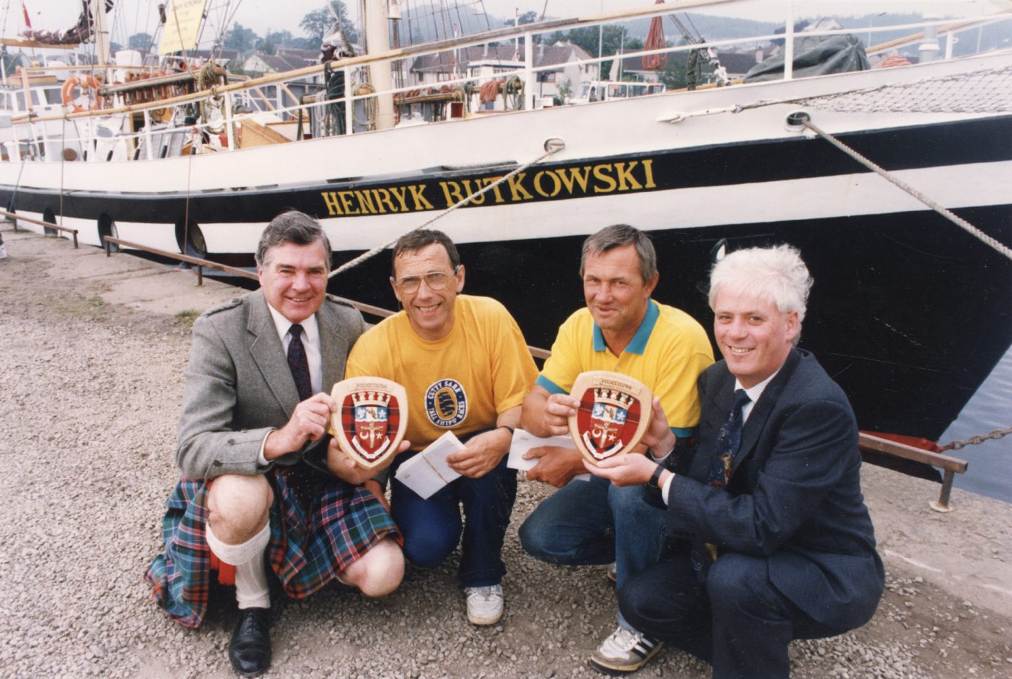 Four men with two coats of arm with a boat behind them