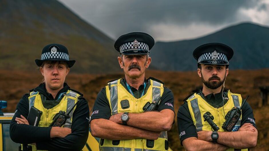 Three police officers, dressed in their uniform, stand with their arms crossed as mountains tower over them in the background.