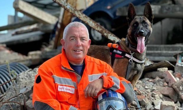 Gary Carroll sitting down on rubble with rescue dog Coorie standing at his shoulder, looking alert