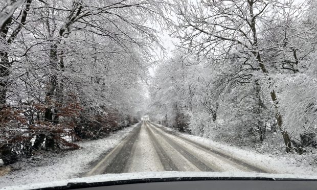 View from the inside of a car driving along a snow covered road lined by trees in Fraserburgh