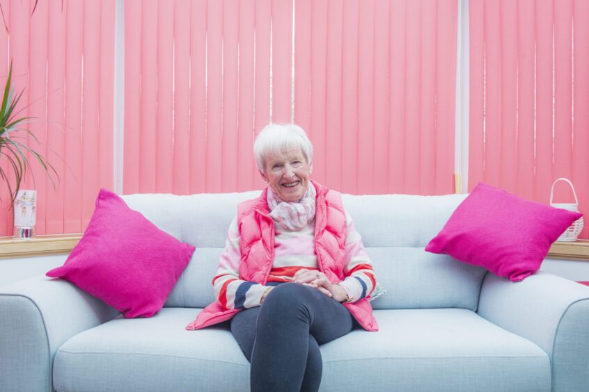 A smiling Liz from Nairn, who has osteoporosis, sits on a blue couch with pink cushions.