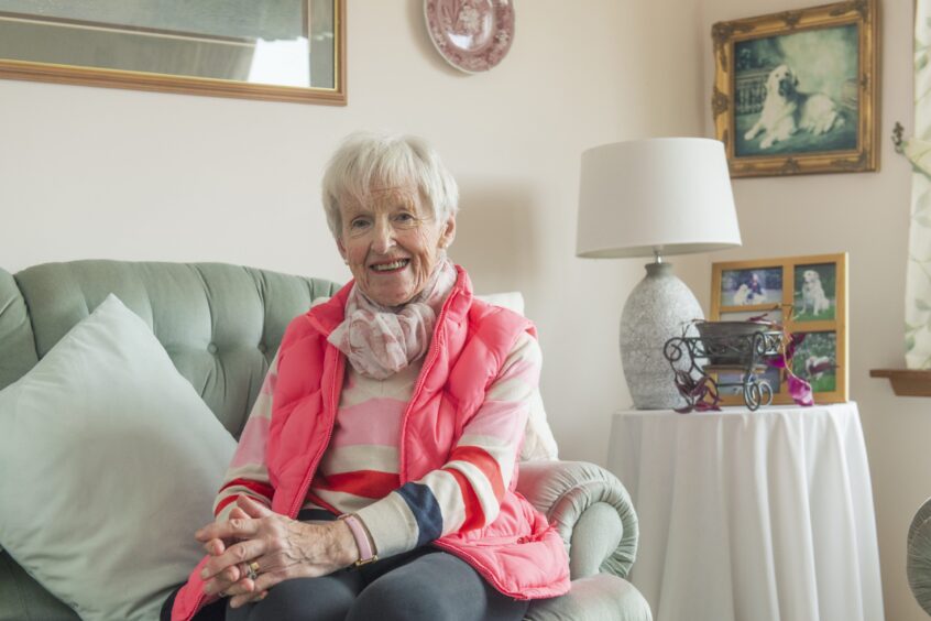 Liz sits on a settee at home in Nairn, smiling and looking at the camera in a pink bodywarmer