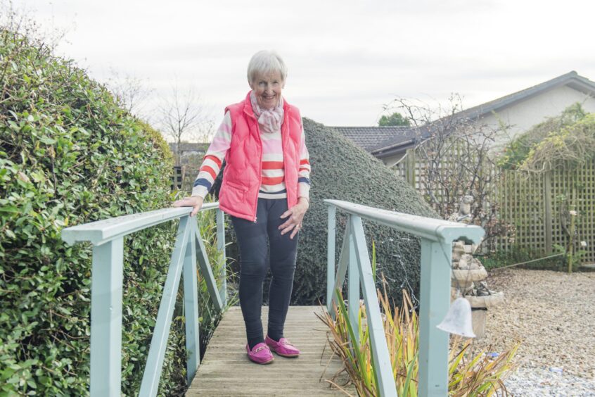 A smiling Liz stands on a wooden bridge in her garden holding on with one hand