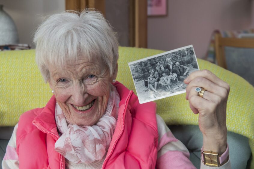 Smiling Liz dressed in pink holds a photo of herself in Millport as an eight-year-old