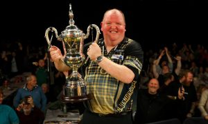 Darts player John Henderson holding the World Seniors Matchplay trophy aloft.