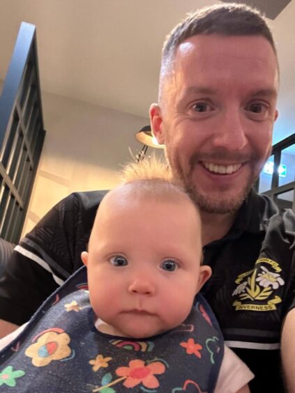 Keith Mason, wearing a black top with the Clachnacuddin FC logo in it, holding his young daughter Sophie wearing a blue flower bib.