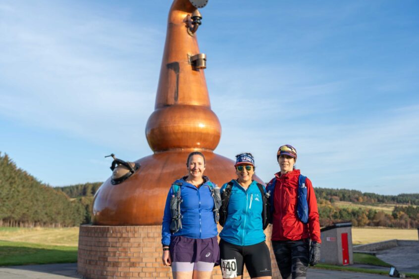 three runners pose for a picture beside a giant, monument in the shape of a whisky still