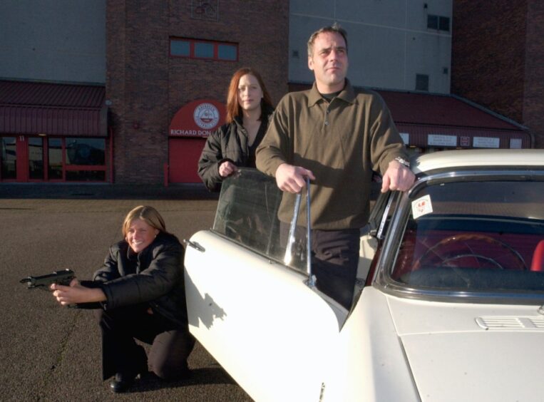 Beside a white car are Aberdeen FC goalkeeper Peter Kjaer with, left, Sarah Duthie, holding a gun, and right, Emma Brand to promote 'The Saint'.