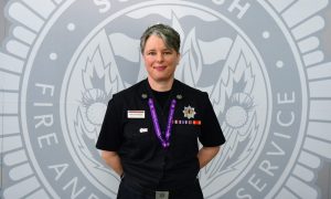 Karla Stevenson stands with her hands behind her back in her black uniform in front of a grey wall with the fire service logo.