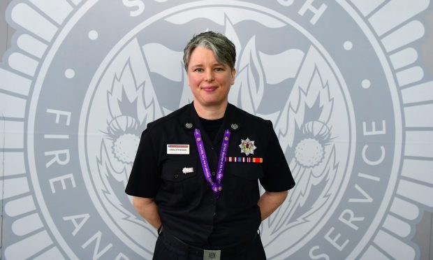 Karla Stevenson stands with her hands behind her back in her black uniform in front of a grey wall with the fire service logo.