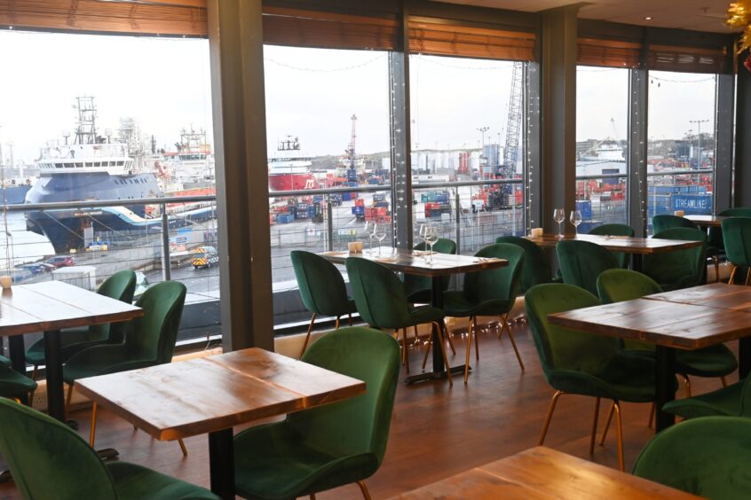 Restaurant interior with green velvet chairs and wooden tables, overlooking a busy harbour with ships, cranes, and shipping containers visible through large floor-to-ceiling windows.