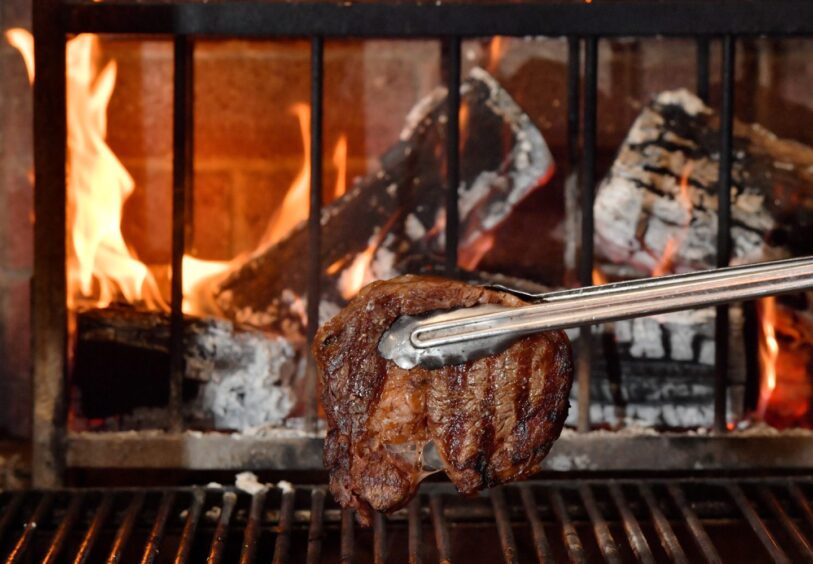 A close-up of a grilled steak held by tongs in front of a wood-burning fire, with glowing embers and flames in the background.