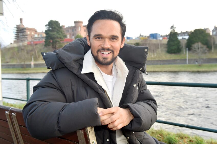 Gareth Gates in a black jacket and white shirt sitting on a bench along the River Ness. 