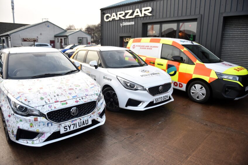 Two MG models and a van wrapped in charity branding sitting side by side outside the car dealership.