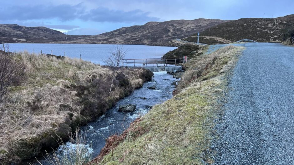 A stream runs by the side of the road, leading to an island loch. The carriageway next to it is of loose stones. 