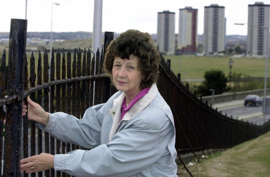 Councillor Jaffrey tests damaged fencing outside the Brig Inn, with city high-rises behind her.