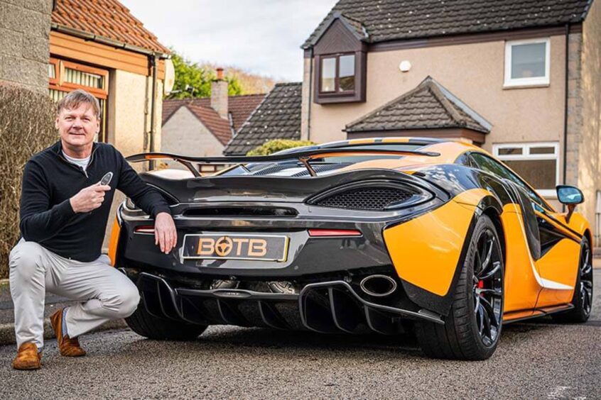 Smiling Aberdeen man David kneels down beside his new black and orange supercar, which is a McLaren he won in a competition