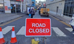 Pedestrians walk behind red road closed sign as vehicles block the street.