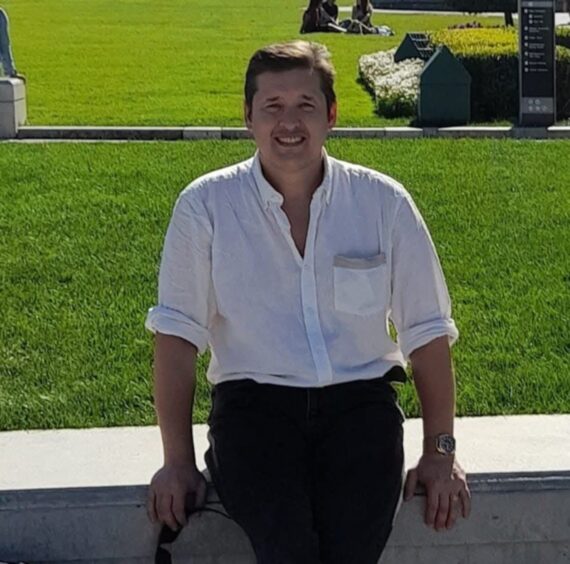 Leslie Sharp leaning against a wall, dressed in trousers and a white shirt.