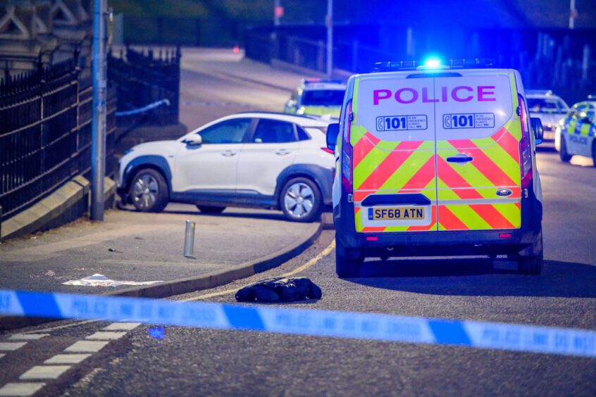 Police van sits behind police tape as a white car sits across the pavement.