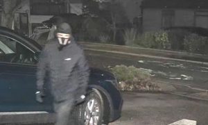 A man dressed all in black, wearing a black hat and a mask appears to enter a driveway.