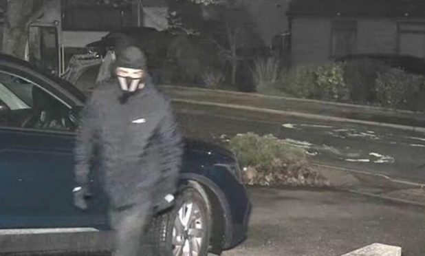 A man dressed all in black, wearing a black hat and a mask appears to enter a driveway.