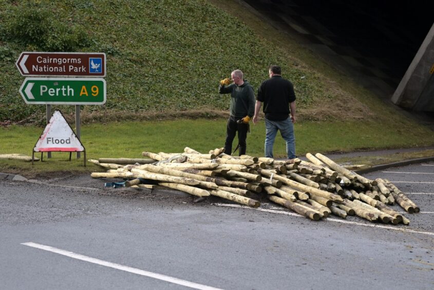 Two men dressed in black in front of a pile of wooden posts.