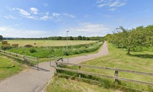 View of footpath from Jubilee Bridge in Nairn with blue spies overhead and fields on both sides.