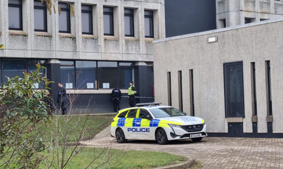 Police car pictures next to concrete wall of Dyce Academy as police stand guard at the main entrance.