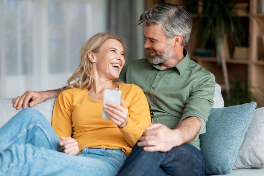 Portrait Of Cheerful Middle Aged Couple Resting With Smartphone On Couch