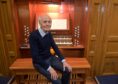 A man in dark trousers and jumper sitting with his hands on his knees in front of a full size church organ