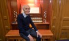 A man in dark trousers and jumper sitting with his hands on his knees in front of a full size church organ