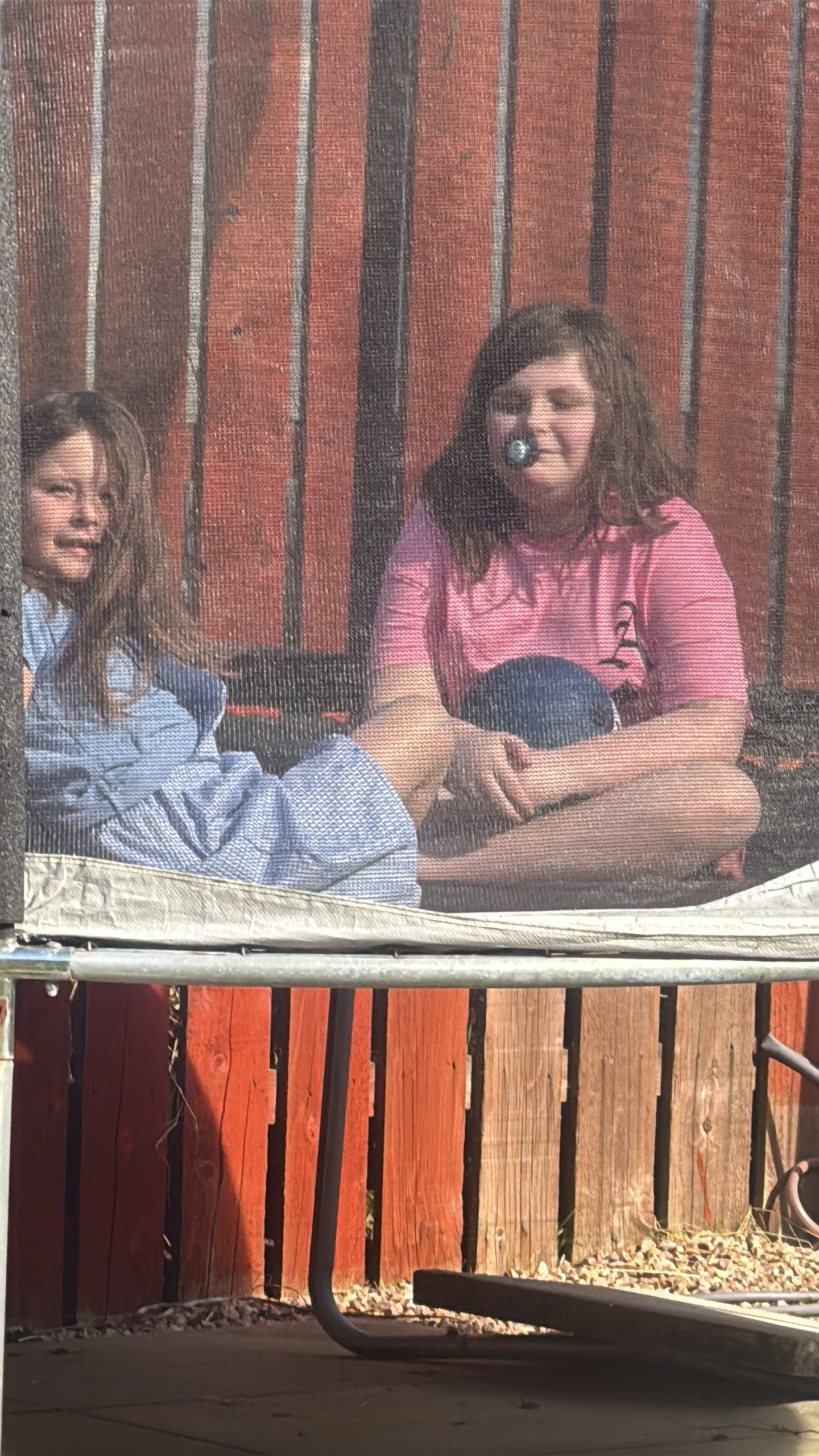 Ariana in a pink T-shirt sitting cross-legged on a trampoline with a black ball in her arms, with her younger sister next to her.