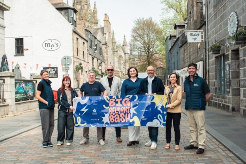 Members of Collective on the Cobbles with a banner to advertise the return of the Big Belmont Bash with Ma Cameron's and the Old School House to the left and Cup and Poldinos to the right.
