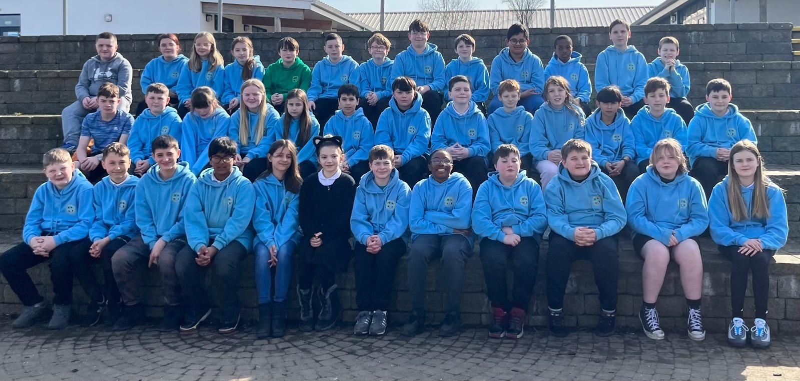 Three rows of pupils in the playground in light blue hoodies.