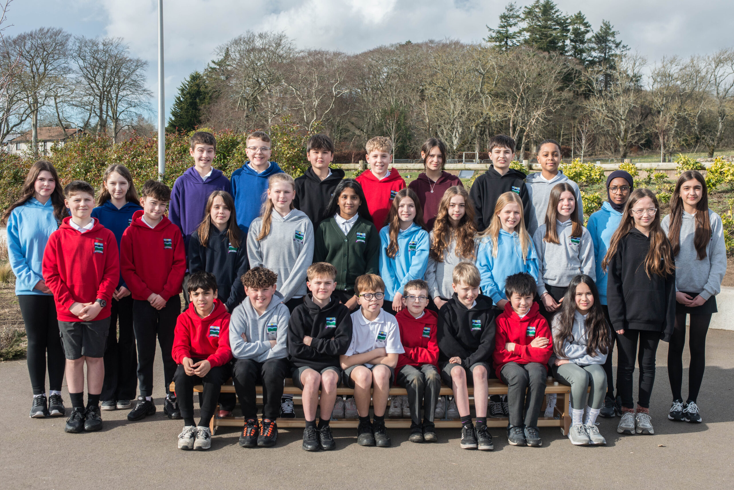 Three rows of pupils on benches outside in various colours of school tops.